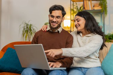 Evdeki oturma odasındaki kanepede dizüstü bilgisayar kullanırken gündelik kıyafetlerini giymiş neşeli çift tartışıyorlar. Mutlu İspanyol aile planlaması dairede online bilet rezervasyonu olduğunu gösteriyor..