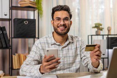 Mutlu Hintli işadamı ofis masasında online alışveriş ödemeleri için banka kartı ve akıllı telefon kullanıyor. İş anlaşması. Gülümseyen adam cep telefonu E-bankacılık uygulama servisiyle serbest alım yapıyor..