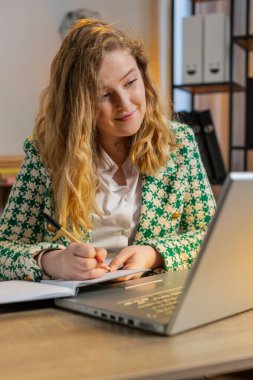 İnternetteki video dersine ya da ofis bilgisayarındaki ofis toplantısına katılırken not alan genç bir iş kadını. Kız serbest çalışan destek servisi web kamerasına bakıyor. Dikey
