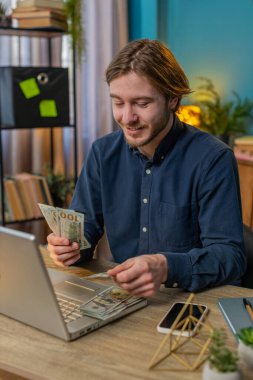 Elinde nakit para banknotlarının hayranı olan bir iş adamı. Başparmaklarını dizüstü bilgisayarına gösteriyor ve işaret ediyor. Kutlama, başarı, internet gelirleri, ofis masasındaki zenginlik. Uzaktan kumandalı. Dikey