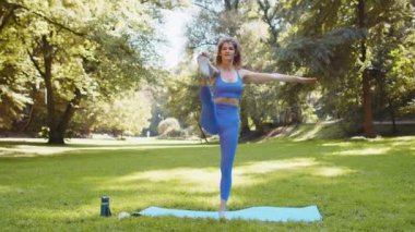 İnce yogalı kadın mavi spor giyer güneşli parkta yoga yapar, bireysel antrenmanlardan zevk alır Ağaç Pozu Vrksasana dengeyi geliştirir ve Utthita Hasta Padangusthasana Uzatılmış Elden Büyük Parmaklı Asana.