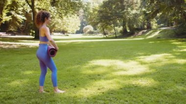 Genç bir kadın parkta çimlere yoga minderi seriyor. Esnek sporcu kadın iyi fiziksel ve zihinsel sağlık için yoga yapıyor. Doğada el kaldırma. Güneşli bir günde dışarıda yoga yap..