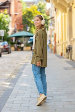 Kablosuz kulaklıklarla gezen mutlu, rahat genç kadın turistin arka görüntüsü açık havada dans eden favori enerjik disko müziğini dinliyor. Güneşli şehir sokağının önünden geçen beyaz bir genç kız..