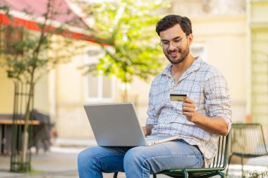 Hintli adam parayı transfer ederken kredi kartı dizüstü bilgisayarı kullanıyor, online alışveriş yapıyor, dışarıdan yemek siparişi veriyor. Şehirde sandalyede oturan genç bir turist..