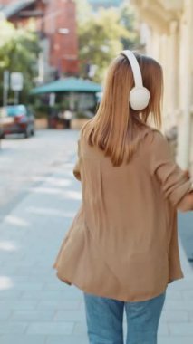 Kablosuz kulaklıklarla gezen mutlu, rahat genç kadın turistin arka görüntüsü açık havada dans eden favori enerjik disko müziğini dinliyor. Güneşli şehir sokağının önünden geçen beyaz bir genç kız..