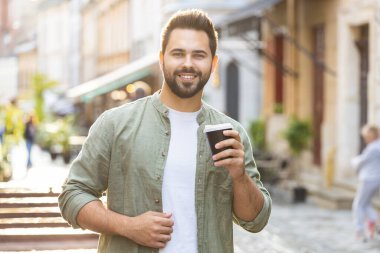 Mutlu sakallı genç adam sabah kahvesinin tadını çıkarıyor sıcak içecek ve dışarıda gülümsüyor. Dinleniyorum, ara veriyorum. Şehirdeki Sunshine Caddesi 'nde yürüyen ve kahve içen bir adam. Dışarıdaki kasaba yaşam tarzları