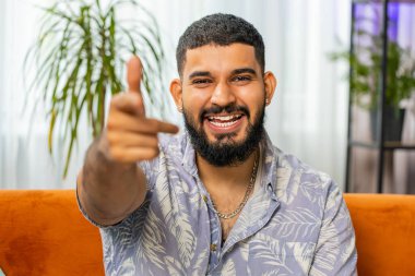 Hey sen! Mutlu Hintli adam heyecanlı bir şekilde gülümsüyor ve kamerayı işaret ediyor, şanslı kazananı seçiyor, muhteşem olduğunu gösteriyor, davet ediyor, onaylıyor. Oturma odasındaki Arap adam kanepede oturuyor.