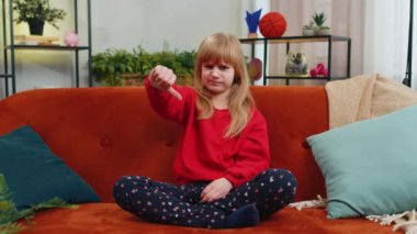 Hoşlanmamak. Mutsuz çocuk kız işaret dilini gösteriyor, hoşnutsuzluğunu dile getiriyor, hoşnutsuzluğunu dile getiriyor, modern apartman dairesinde tatminsiz kötü işler yapıyor. Oturma odasındaki kanepede oturan hoşnutsuz genç çocuk.