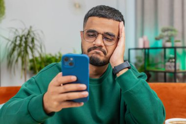 Hüzünlü Arap adam akıllı telefon daktilo kullanıyor, beklenmedik piyango sonuçlarını kaybediyor, kötü haber, servet kaybı, son teslim tarihi, virüs. Masadaki ofisteki genç Hintli adam.