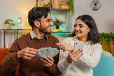 Happy Indian couple holding pointing on money cash and smartphone planning discussing investment together sitting on sofa in living room at home. Successful Arabian family on couch in apartment.