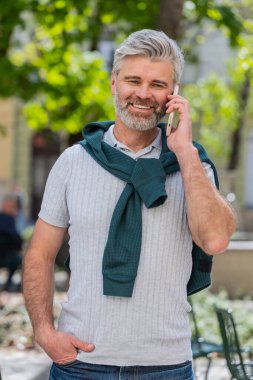 Sakallı, olgun bir turist akıllı telefonla konuşuyor. Şehir caddesinde beklenmedik bir şekilde iyi haber dedikodusu yapıyor. Şehir yaşam tarzları açık havada.
