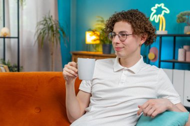 Mutlu genç adam içecek kokusu alıyor sıcak kahve, dinleniyor, dinleniyor. Gülümseyen beyaz adam aroma çayını tek başına içmeyi sever. Oturma odasındaki kanepede oturur..