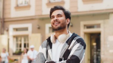 Kablosuz kulaklıklı, boynuna bakıp gülümseyen gündelik kıyafetlerle düşünceli genç bir turist. Şehir caddesinde dikilen mutlu, pozitif İspanyol gezgin adam. Kasaba yaşam tarzları.