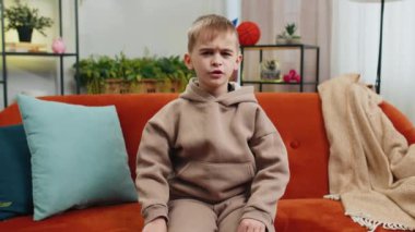 Hoşlanmamak. Üzgün çocuk işaret dilini gösteriyor, hoşnutsuzluğunu ifade ediyor, hoşnutsuzluğunu dile getiriyor, modern ev dairelerinde tatminsiz kötü işler yapıyor. Oturma odasındaki kanepede oturan hoşnutsuz genç çocuk.