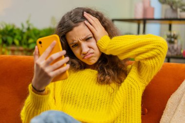 Küçük kız akıllı telefon kullanıyor, oyun oynuyor, beklenmedik piyango sonuçlarını kaybediyor, kötü haber, servet kaybı, başarısız eğitim sınavı. Oturma odasındaki genç kız kanepede oturuyor.