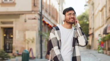 Kablosuz kulaklıklı mutlu Arap genç turist açık havada akıllı telefondan favori enerjik disko müziğini dinliyor. Şehir sokağının yanından geçen Hintli turist..