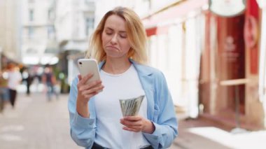 Mutlu, genç, sarışın bir kadın nakit para sayıyor, akıllı telefon hesap makinesi uygulaması kullanıyor, internetten hediye ve yemek siparişi vermeyi planlıyor, dışarıda otel odası ayırtıyor. Şehir caddesinde duran kadın.