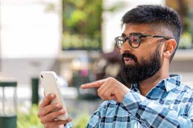Hintli sakallı adam akıllı telefon kullanıyor sosyal medya uygulamasında mesaj yazıyor, internette sörf yapıyor, dinleniyor, dışarıda mola veriyor. Şehir caddesinde duran İspanyol adam. Yatay