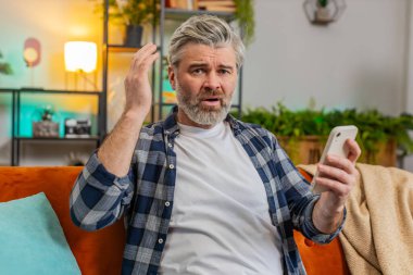 Üzgün, olgun bir adamın elinde akıllı telefonuyla oturup olumsuz mesaj okuması oturma odasında kanepede oturmaktan rahatsız ediyor. Beyaz, yaşlı, yaşlı bir adam dairede alet sorunu yaşıyor. Uygulama çökme konsepti.