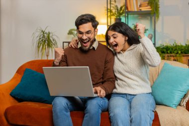 Evdeki oturma odasındaki kanepede e-posta kazanan piyango ikramiyesine şaşırmış, gündelik giysiler içinde neşeli genç çift. Mutlu Hint ailesi dairede yumruklarını sıkıyor.