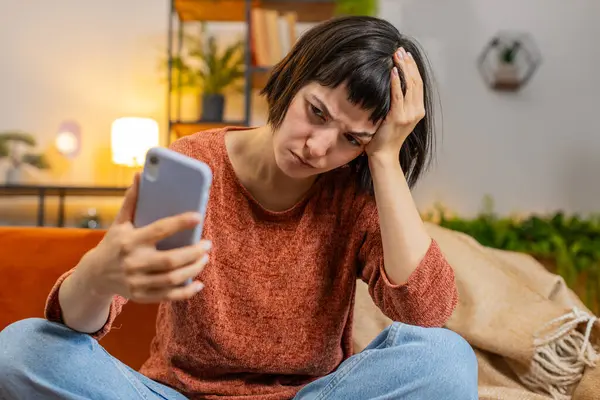 stock image Upset displeased woman with short hair using smartphone sitting on sofa in living room at home. Young Hispanic girl in casual clothes loses becoming surprised by sudden lottery results or bad news.