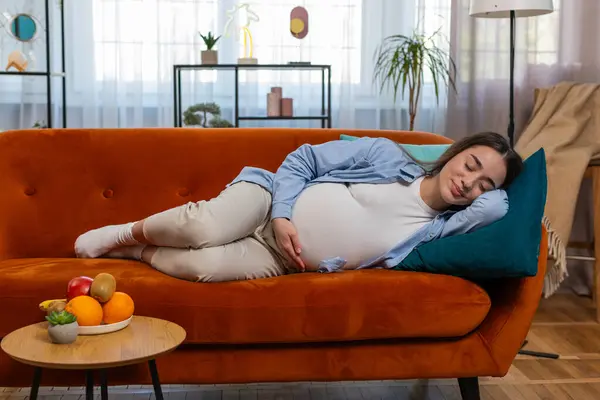 stock image Smiling young pregnant woman stroking big belly while sleeping on comfortable cozy sofa dreaming in living home room. Happy future mother lady in casual clothes relaxing on couch in modern apartment