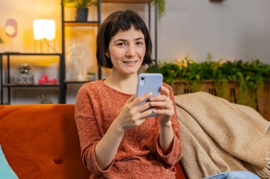 Akıllı telefondan düşünen ve mesaj atan mutlu genç bir kadın. Kanepede oturan ve cep telefonu kullanan İspanyol kız evdeki oturma odasında kameraya bakıyor. Dairede internetten film izleyen bir kadın..