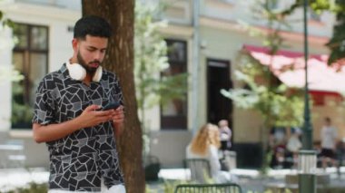 Mutlu genç Hintli adam kablosuz kulaklık takıyor, açık havada akıllı telefondan en sevilen enerjik müziği dinliyor. Sakallı adam şehir parkının yanından geçiyor. Yaşam Tarzları.