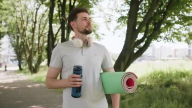 Spor antrenmanından sonra yoga minderiyle atletik spor koşucusu şişeden su içiyor. Parkta yürüyen kardiyo antrenmanı. Genç koşucu sağlıklı yaşam tarzını seviyor. Aktif sporcu aktiviteleri.
