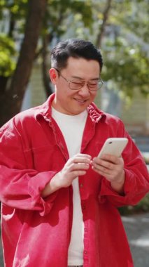 Mutlu bir Asyalı orta yaşlı adam akıllı telefon kullanıyor internette mesajlar yazıyor, işi bitiriyor, dışarıdaki kameralara bakıyor. Şehir sokağındaki Çinli adam. Kasaba yaşam tarzları. Dikey