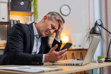 Üzgün genç işadamı, ofis masasında akıllı telefondan kötü haberler okuyor. Resmi takım elbiseli üzgün erkek işveren hoş olmayan tebligattan rahatsız, internetteki bilgiden şok olmuş, mesajla depresyona girmiş..