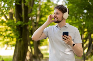 Sporcu spor koşucusu adam yürüyor ve akıllı telefon kullanıyor kablosuz kulaklıkla müzik dinliyor. Şehir parkında egzersiz kardiyosu. Genç koşucu sağlıklı yaşam tarzını seviyor..