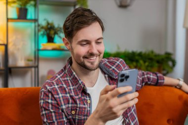 Sıradan kıyafetler içinde mutlu sakallı bir adam akıllı telefondan düşünüyor ve mesajlaşıyor. Beyaz adam kanepede oturuyor ve evdeki oturma odasında cep telefonu kullanıyor. Erkek, dairede online film izliyor..