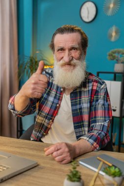 Gibi. Gülümseyen kıdemli adam kameraya onaylayıcı bir şekilde bakıyor. İki başparmak işareti gösteriyor. İyi haber olumlu geri bildirim. Ev ofis masasında dizüstü bilgisayarı olan mutlu büyükbaba. Dikey