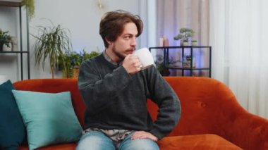 Çekici, gülümseyen, sakallı bir adam bir fincan sıcak kahve ya da bitki çayı içerken sabah evde oturma odasındaki kanepede oturuyor. Kafkasyalı genç yetişkin adam tek başına rahatça dinleniyor.