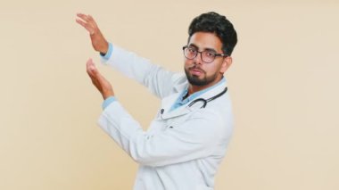 Hintli genç doktor kardiyolog etrafa para saçarak vakit kaybettiğini gösteriyor. Daha fazla bahşiş, büyük kar, kazanma, ödeme satın alma. Arap eczacı, bej arka planda izole edilmiş.