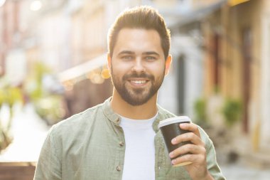 Mutlu sakallı genç adam sabah kahvesinin tadını çıkarıyor sıcak içecek ve dışarıda gülümsüyor. Dinleniyorum, ara veriyorum. Şehirdeki Sunshine Caddesi 'nde yürüyen ve kahve içen bir adam. Dışarıdaki kasaba yaşam tarzları