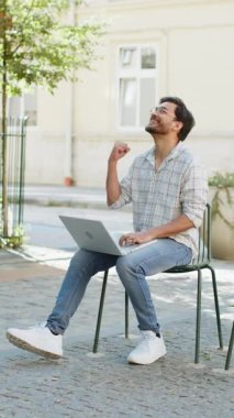 Laptop kullanarak kutlama yapan yakışıklı genç adam iyi haber kazandı, piyango zaferi kazandı, açık havada internetten hediye verdi. Kafe terasında oturan mutlu Hintli turist. Kasaba yaşam tarzları.