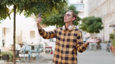 Neşeli blog yazarı akıllı telefondan selfie çekiyor abonelerle ya da aile dostlarıyla görüntülü iletişim kuruyor sosyal medya vlog 'u için hikayeler kaydediyor, el sallıyor. Şehir sokağındaki adam..