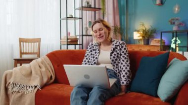 Aşırı mutlu genç bir kadın bilgisayarla arkadaşlarıyla konuşuyor. Gülümse, el salla, ekranda iyi haberler var. Mutlu beyaz kız evde kanepede oturup sanal sohbetten zevk alıyor..