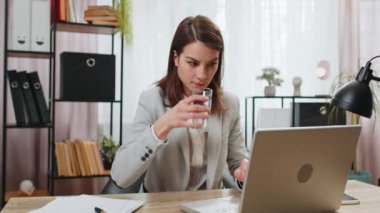 İşyerindeki masa başında oturmuş laptopla çalışırken su içen genç bir iş kadını. Kadın çalışan serbest çalışan, bilgisayar ekranlı internette sörf yapıyor. İyi alışkanlıklar.
