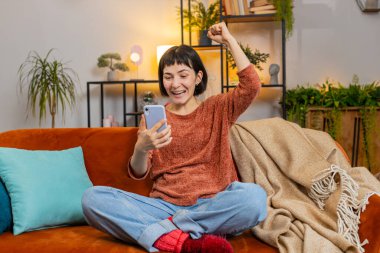 Şaşırmış mutlu kadın akıllı telefon piyangosuna bakıyor evdeki kanepede oturuyor. Heyecanlı Kafkasyalı kız dairede zafer kutlaması yapıyor yumruklarını sıkıyor. Mutlu bayan çevrimiçi oyun oynuyor