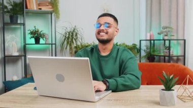 Şaşırmış Hintli genç adam dizüstü bilgisayar kullanıyor, iyi haber mesajı alıyor ani zaferin şokuyla piyango kazanma ikramiyesini kutluyor evdeki dairede online alışveriş oyunu oynuyor.