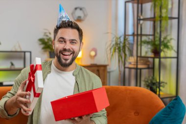Mutlu genç adam doğum günü şapkası takıyor ve elinde kurdeleyle kutluyor. Beyaz tenli esmer erkek, parti açılışını kutluyor. Kanepede, toplantı odasında hediyeler sunuyor..