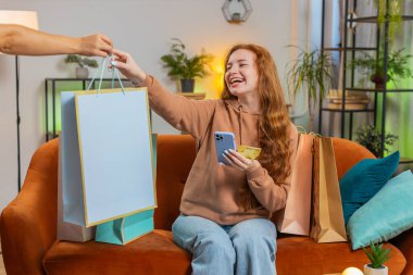 Alışveriş meraklısı kızıl saçlı mutlu bir kadın alışveriş torbalarıyla evde oturuyor online sipariş veriyor, kredi kartı ile ödeme yapıyor. Evdeki koltukta oturan heyecanlı kadın...