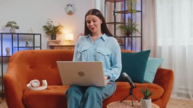 Happy blogger woman recording podcast at desk, giving workshop, webinar. Young Caucasian girl speaking at microphone and laptop webcam, making video call, discussing at home living room sits on couch