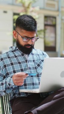 Kredi kartı ve dizüstü bilgisayar kullanan Hintli sakallı bir adam internetten alışveriş siparişi verirken açık havada internetten sipariş veriyor. Şehir sokağındaki İspanyol adam.