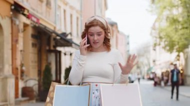 Alışveriş sonrası alışveriş yapan mutlu kızıl saçlı kadın açık havada hediyelerle dolu poşetlerle. Şehir caddesinde yürüyen turist kız akıllı telefondan uzaktan konuşma yapıyor.