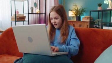 Bilgisayarlı daktilo kullanan mutlu bir kız çocuğu büyük bir sürpriz buldu, olumlu haberler, online maç kazancını kutladı. Oturma odasında kanepede oturan neşeli kız çocuk..