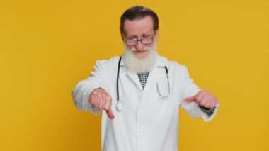 Hoşlanmamak. Mutsuz yaşlı doktor kardiyolog. Başparmak işareti gösteriyor. Onaylanmamış olumsuz geri bildirim. Eczacı dedesi sarı arka planda izole edilmiş.
